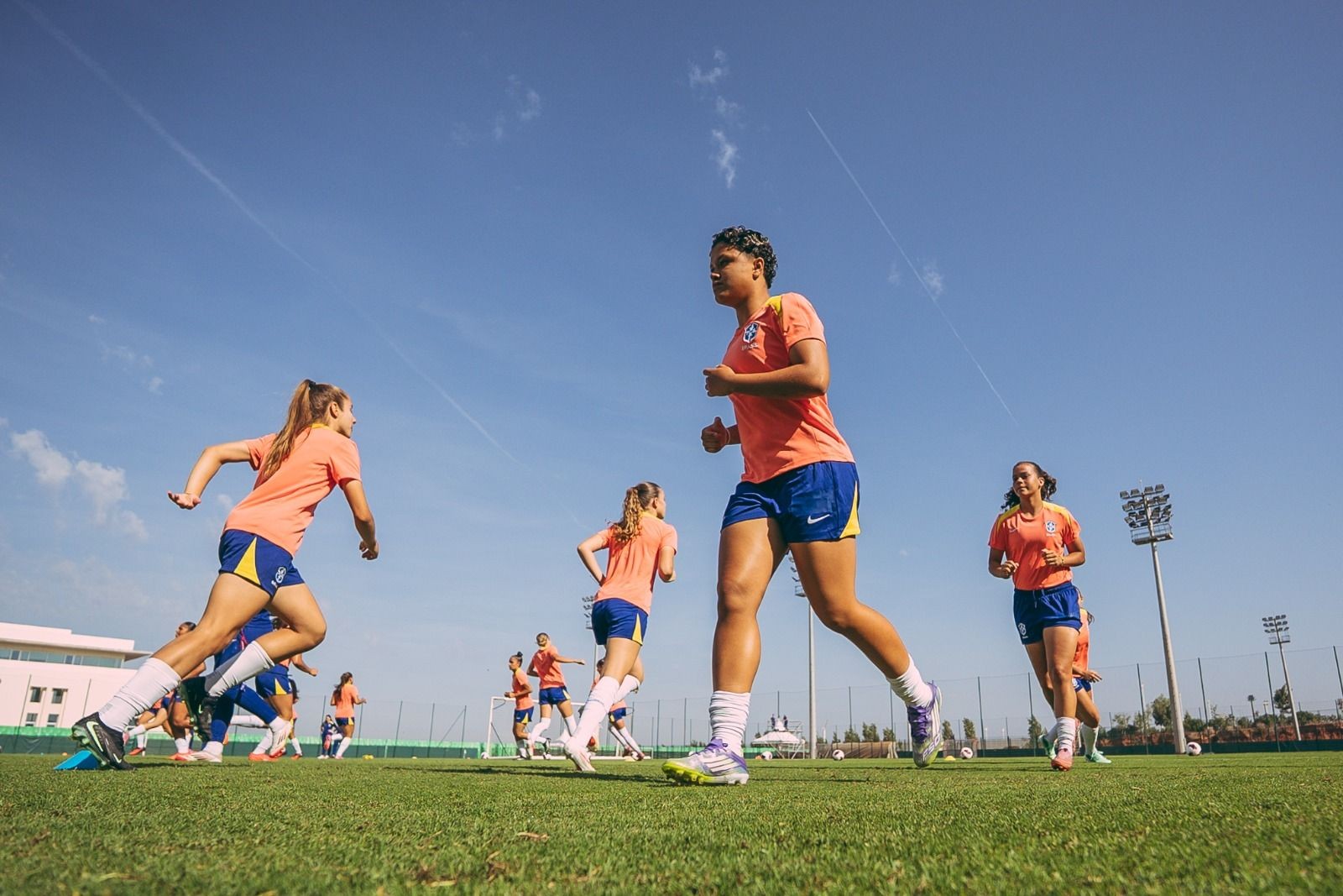 Jogadoras de futebol feminino correndo em campo de grama verde sob céu azul claro. Uma mulher de camiseta laranja e shorts azuis com detalhes amarelos corre em primeiro plano, à direita. Outras jogadoras, com uniformes semelhantes, correm ao fundo, algumas com o cabelo preso em rabo de cavalo. Há um prédio branco à esquerda e torres de iluminação à direita. A cena transmite energia e movimento.