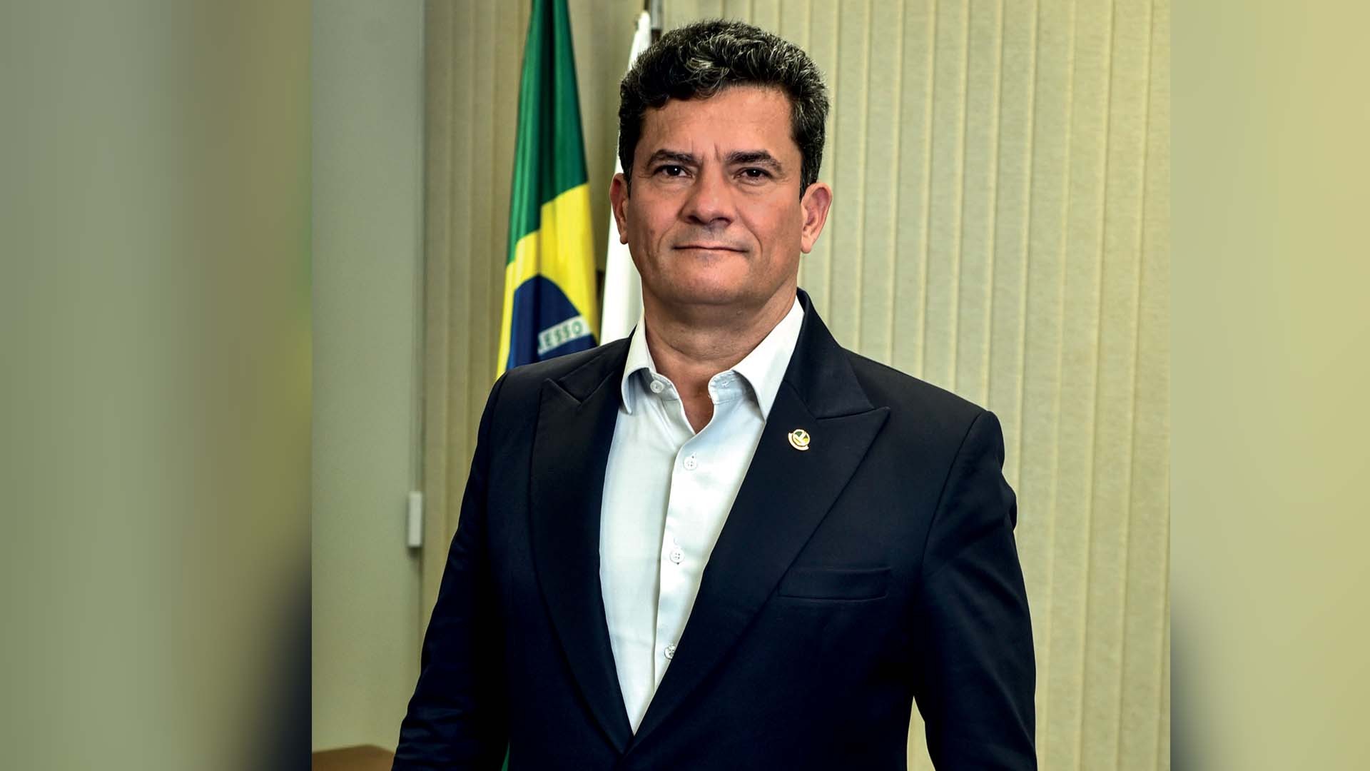 Homem de pele clara, cabelo escuro e curto, vestindo blazer azul-marinho com broche dourado e camisa branca desabotoada, em pé, olhando para frente com expressão séria. Ao fundo, uma bandeira do Brasil e uma parede bege
