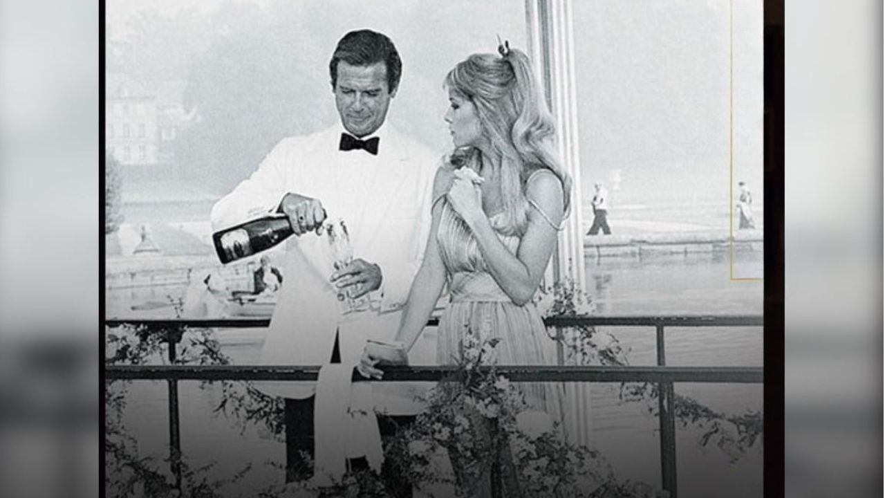 Roger Moore, de smoking branco e gravata borboleta preta, serve champanhe para Barbara Bach, de vestido claro e cabelos loiros soltos, em uma varanda com vista para um lago e construções ao fundo