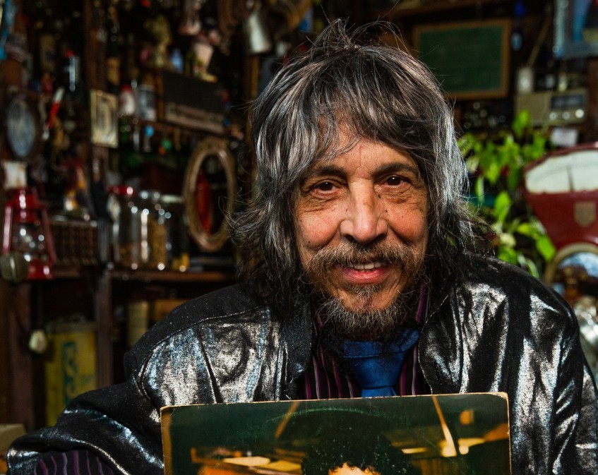 Homem de pele morena, cabelos grisalhos longos e barba, sorri levemente, vestindo jaqueta preta brilhante e gravata azul, segurando um disco de vinil com capa escura. Ao fundo, prateleiras com objetos antigos e plantas