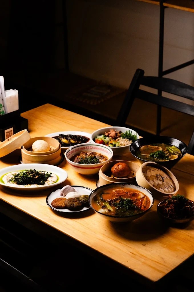 Mesa de madeira clara com diversos pratos asi&aacute;ticos: sopas, saladas, bolinhos em cestas de bambu, e outros pratos com molhos escuros e vegetais verdes, sob ilumina&ccedil;&atilde;o focada